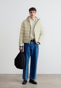 Νεαρός άνδρας που φοράει μπεζ φουσκωτό μπουφάν, ανοιχτό πουλόβερ πόλο, μπλε τζιν, μαύρα παπούτσια, κρατώντας μια μαύρη τσάντα, στέκεται μπροστά σε απλή ανοιχτόχρωμη τοίχο.