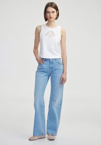 Top blanco sin mangas con detalle de encaje floral, combinado con jeans de pierna ancha azul claro con lunares blancos. Zapatos de color neutro.