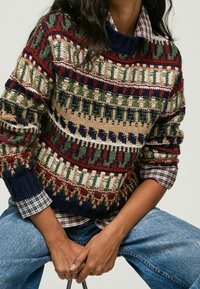 Pull en tricot présentant des motifs multicolores en rouge, vert, bleu marine et crème, avec un col montant et des poignets côtelés. Associé à une chemise à carreaux.