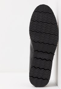 Svart gummisåle med zigzag-mønster, med en liten størrelsesindikator på siden. Glatt overflate med definerte kanter.