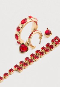 Smykkesett i gullfarge med røde edelstener, inkludert en ring med hjerteanheng, hoop-øreringer og et armbånd med sammenknyttede steiner.
