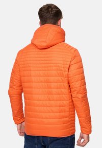 Oranssi tikattu puffertakki, jossa on huppu, hieno pinta ja vaakasaumotus. Pitkät hihat ja vyötäröllä hyvin istuva malli.