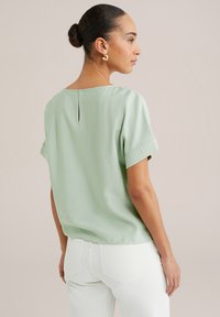 Hele roheline lühikeste varrukatega topp, millel on ümar kaelus ja väike lõhe seljaosas, valmistatud pehmest ja tekstuurist kangast. Kombineeritud valgete pükstega.