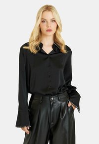 Blusa de satén negra con cuello puntiagudo adornado con tachuelas, cierre de botones y mangas acampanadas con puños con tachuelas, acompañada de pantalones de cuero.