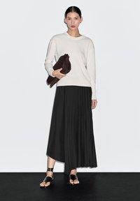 Lichtgrijze trui, zwarte geplooide rok en zwarte sandalen. Het model houdt een donkerbruine clutch vast, tegen een plain witte achtergrond.