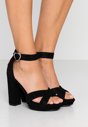 Black suede block-heeled sandals feature a cross design on the toe, ankle strap with a heart-shaped buckle, and a textured finish.
