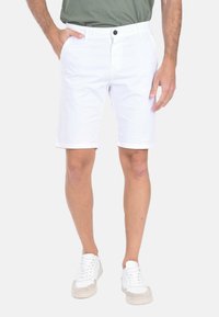 Pantaloni corti bianchi in cotone con taglio dritto, dotati di chiusura con bottone e tasche laterali. Indossati con una camicia verde e sneakers bianche.