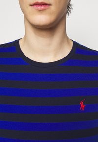 Blau- und marineblaue gestreifte T-Shirt mit Rundhalsausschnitt. Mit einem kleinen roten bestickten Logo auf der Brust. Hergestellt aus Baumwollmaterial.