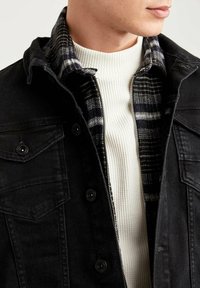 Zwarte denimjack met drukknopen, met een ritssluiting in een geruite flanellen voering. Daaronder is een textuur wit ribbeltrui zichtbaar.