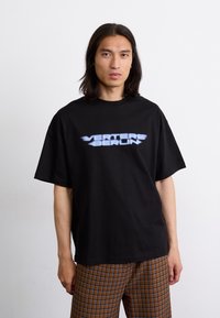 Czarny oversize t-shirt z bawełny, z gradientowym logo "VERTERE BERLIN" w jasnoniebieskim kolorze na piersi. Model w kratkowanych spodniach.