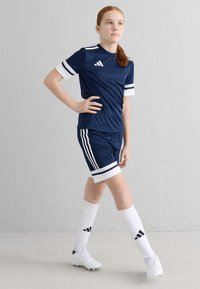 Tumši zils sporta kreklis un šorti ar baltām akcentēm; trīs svītru dizains uz piedurknēm un šortiem. Baltas zeķes un sporta apavi papildina apģērbu.