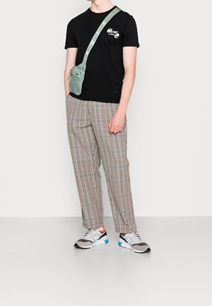 Persona che indossa una maglietta nera con grafica, pantaloni beige a quadri, scarpe da ginnastica grigie e una borsa a tracolla verde con una tracolla con logo.