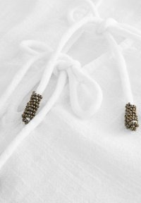 Witte stoffen stropdas met textuuraccenten van bronzen kralen, die een glad oppervlak en ingewikkelde details bij de knoop tonen.