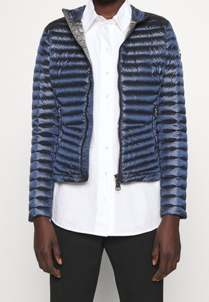 Persoon draagt een marineblauw doorgestikt jack, open over een witte blouse en zwarte broek, staand tegen een effen achtergrond.