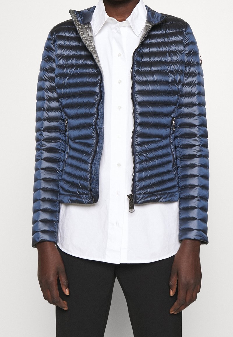 Osoba ubrana w granatową pikowaną kurtkę rozpiętą na białą koszulę z guzikami i czarne spodnie, stojąca na jednolitym tle.