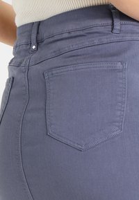 Jeans de mezclilla gris oscuro con un diseño ajustado, que cuentan con un bolsillo trasero y costuras visibles a lo largo de las costuras. Textura suave.