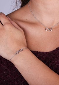 Zilveren infinity halsketting en armband, met een glad ontwerp en een delicate ketting, gedragen op een hand tegen een gestructureerd, donker kledingstuk.