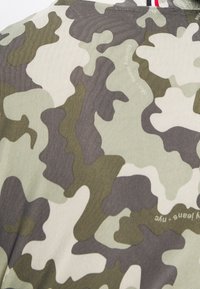 Kamouflage-mönstrad tyg i nyanser av grönt och krämfärgat, med en texturerad yta och oregelbundna former genom hela.