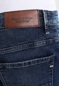 Džíny z tmavě praného denimu, s hnědým koženým záplatou s raženým logem a textem. Obsahují zadní kapsu a prošité detaily.