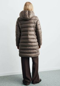 Abrigo tipo puff en marrón apagado, con capucha y diseño acolchado horizontal. Llevado sobre pantalones marrones oscuros, con una textura suave.