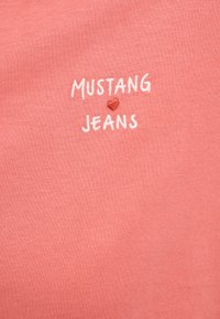 Tissu corail avec texte blanc brodé "MUSTANG" et "JEANS", séparés par un petit cœur rouge au centre.