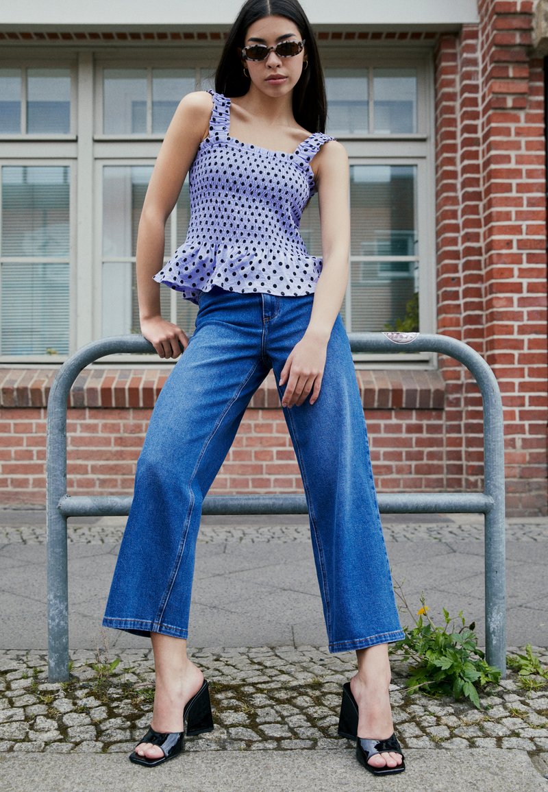 Top de peplum roxo com bolinhas brancas e alças com babados, combinado com jeans azuis de cintura alta e perna larga e sapatos de salto aberto pretos.