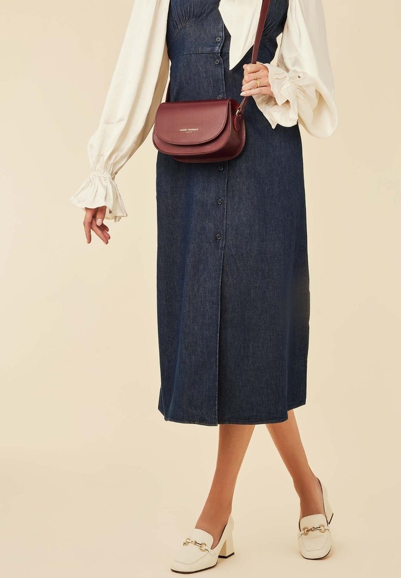 Midi-Denimkleid mit Knopfleiste, langen Ärmeln und weißen Rüschensäumen; burgunderfarbene Leder-Umhängetasche; weiße Absatzslipper.