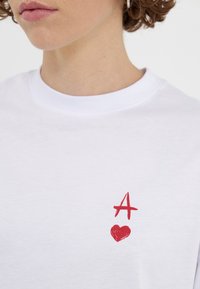 Vit bomull t-shirt med en röd handritad bokstav "A" och hjärtgrafik på vänster sida nära kragen. Enkelt mönster, rund hals.