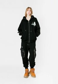 Joven de pie que lleva puesta una sudadera negra con cremallera y un gráfico, pantalones cargo negros y botas de cordones color beige, contra un fondo blanco liso.