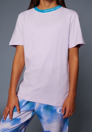 Person iført en ensfarvet lys lilla T-shirt med blå krave tekst "Calvin Klein Jeans" og blå og hvide tie-dye bukser mod en grå baggrund.