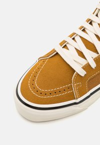 Brązowy zamszowy trampek z białymi sznurówkami, teksturowaną cholewką zamszową, czarnym obramowaniem i perforacjami na nosku.