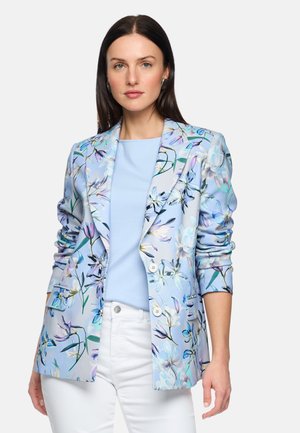 Frau trägt ein hellblaues, floral gemustertes Sakko über einem pastellblauen Oberteil und weißen Hosen, steht vor einem schlichten Hintergrund.