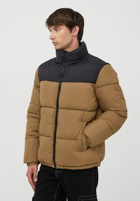 Giacca imbottita con una sezione superiore nera e una sezione inferiore marrone, dotata di un colletto alto, chiusura con zip e polsini elasticizzati per il calore.