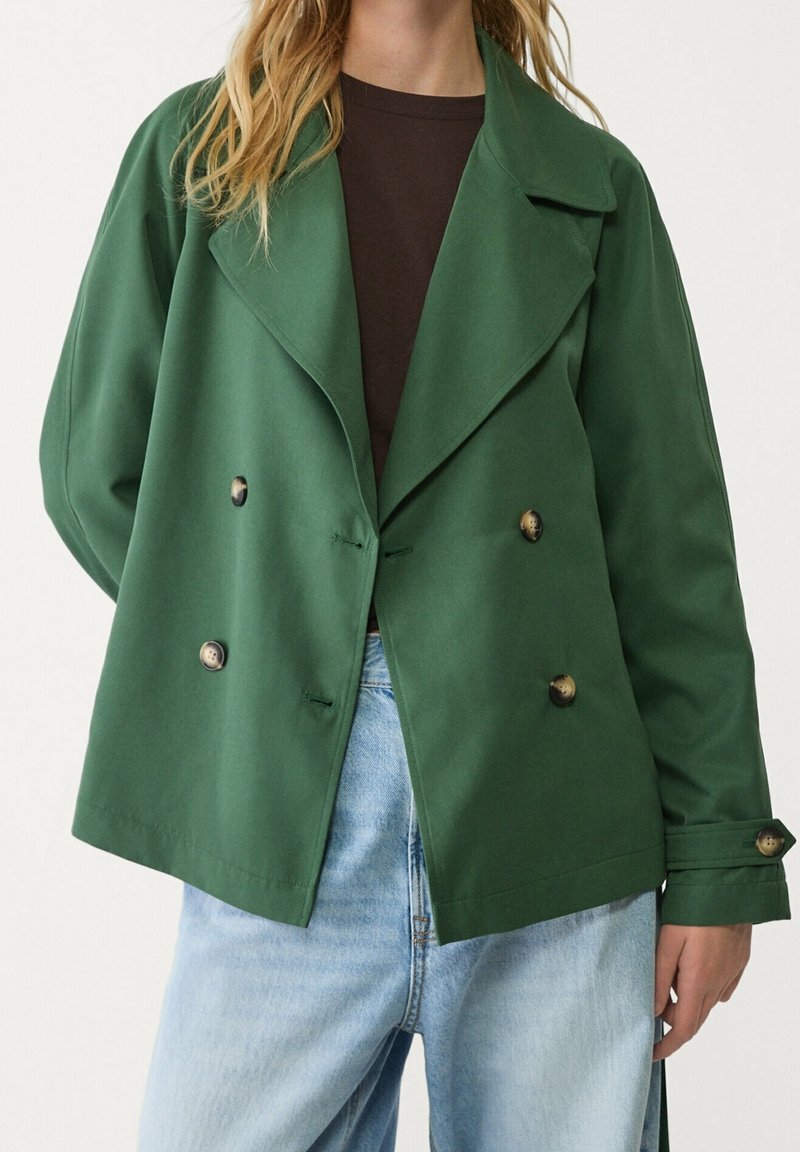 Grøn dobbeltradet jakke lavet af glat stof med store klapper og to mørke knapper. Båret over en mørk skjorte og parret med lyse jeans.