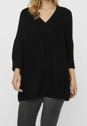 Femme portant un pull ample noir à col en V avec un tricot texturé et des manches trois-quarts, associé à un jean gris foncé.