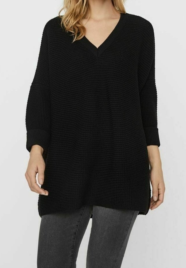 Femme portant un pull ample noir à col en V avec un tricot texturé et des manches trois-quarts, associé à un jean gris foncé.