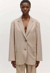 Beige op maat gemaakte blazer met een geknipt revers, een enkele borstzak, twee voorzakken en gouden knopen. Soepele stof en een relaxte pasvorm.