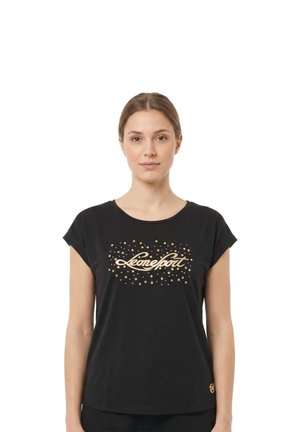 Une femme se tient devant un fond blanc, vêtue d’un tee-shirt noir avec une inscription dorée et un motif à pois.
