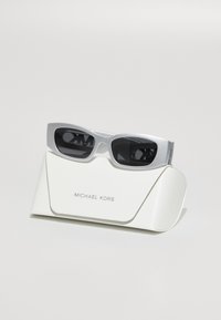Ochelari de soare dreptunghiulari argintii cu lentile întunecate, așezați pe un toc alb texturat cu "MICHAEL KORS" embosat în argint.