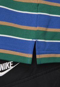 Gros plan sur une chemise à rayures vertes, bleues, beiges et blanches avec une petite fente latérale, portée sur un pantalon noir avec un logo Nike blanc visible.