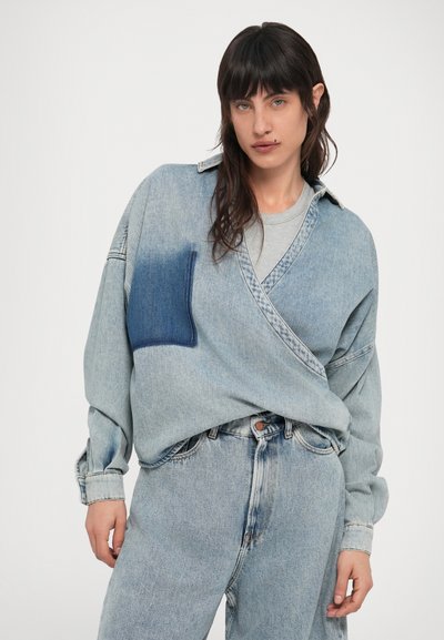 Νεαρό άτομο με σκούρα μαλλιά που φοράει ξεβαμμένο τζιν παντελόνι και υπερμεγέθη τζιν πουκάμισο με σκούρο μπλε μπαλώμα στο στήθος, στέκεται μπροστά σε λευκό φόντο.