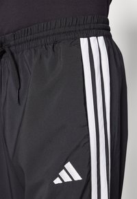 Fekete sportnadrág, húzózsinóros derékkal, fehér oldalsávokkal és hímzett logóval az alsó lábszáron. Simító, könnyű szövet.