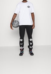 Vit t-shirt med tryckt logotyp, svarta sweatpants med olika laglogotyper och flerskiftande sneakers som håller en basketboll.