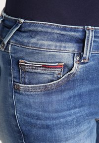 Primer plano de unos vaqueros de mezclilla azul con un pequeño detalle de tela doblado en rojo, blanco y marino dentro del bolsillo delantero de una persona que lleva una camiseta oscura.