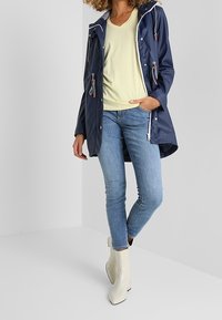 Giacca impermeabile blu navy con cappuccio, dotata di lacci a strisce, chiusura con zip, indossata sopra una maglietta a V gialla chiara e jeans blu.