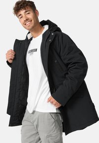 Parka noire avec une capuche en fourrure, boutons-pression et poches latérales. Portée sur un t-shirt blanc et un pantalon cargo gris.