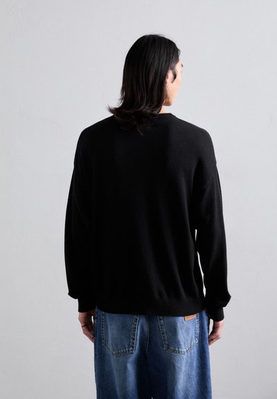 Schwarzer Strickpullover mit rundem Ausschnitt und fallenden Schultern, kombiniert mit hellblauen, weit geschnittenen Jeans. Weiche Textur, minimalistisches Design.