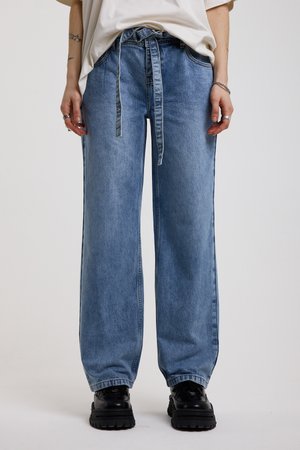 Άτομο που φοράει ανοιχτό μπλε τζιν με φαρδιά μπατζάκια και δεμένη ζώνη στη μέση, λευκό πουκάμισο, μαύρα chunky παπούτσια και ασημένια δαχτυλίδια και βραχιόλια στα χέρια.