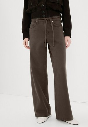 Pantalones marrones de pierna recta con un cinturón de tela anudado, combinados con una chaqueta negra abotonada y zapatos blancos sobre un fondo liso.