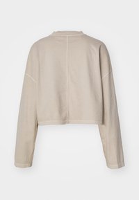 Beige cropped huppari, jossa on pitkät hihat, pyöreä kaula-aukko ja näkyvä sauma, joka kulkee alas keskiselästä.